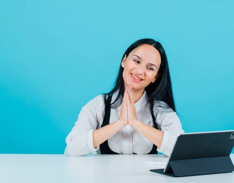 Smiling blogger girl is holding hands together by posing to tablet camera  on blue background